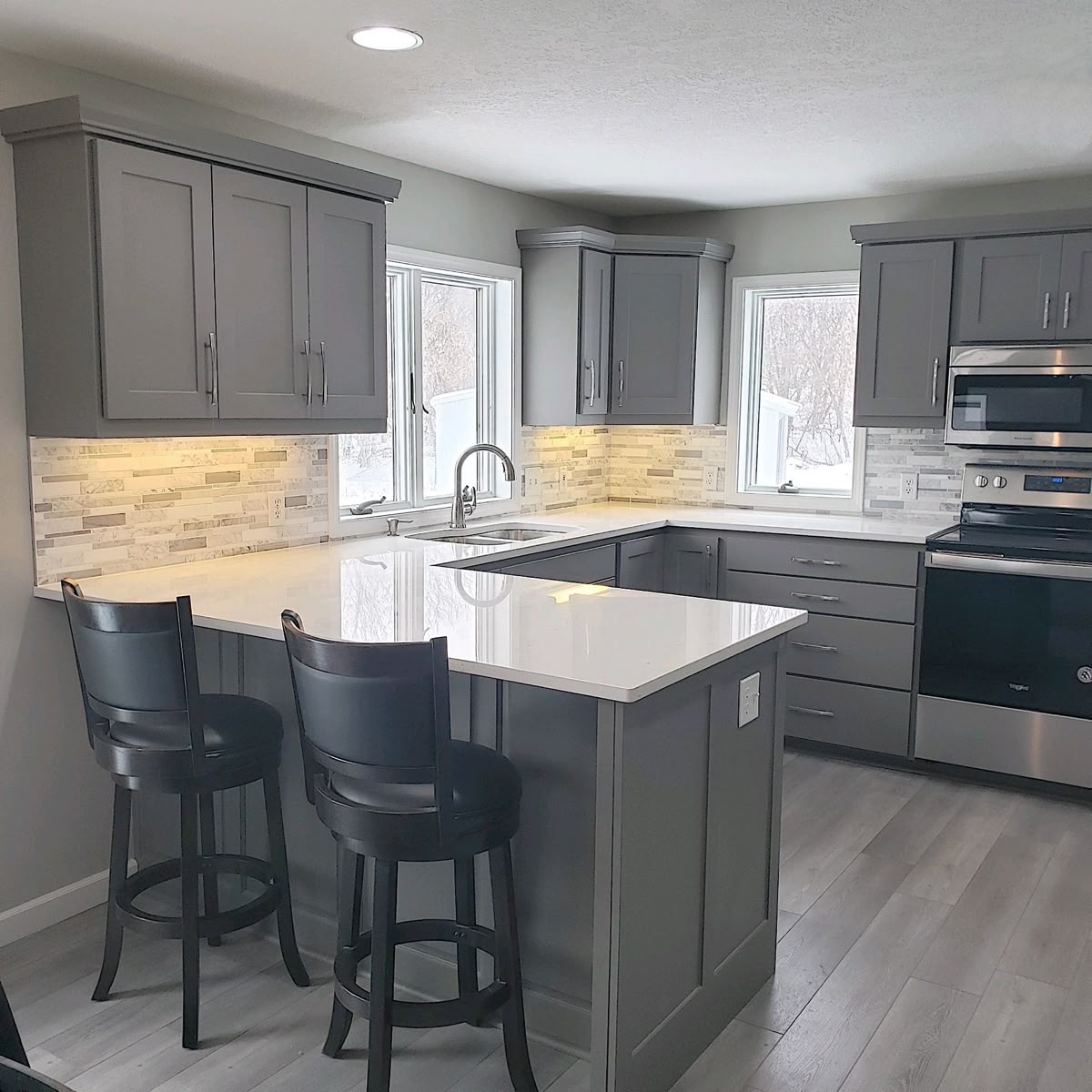 Modern kitchen with white upper cabinets, dark lower cabinets, stainless steel appliances, herringbone wood floor, and a large marble countertop island with two stools.