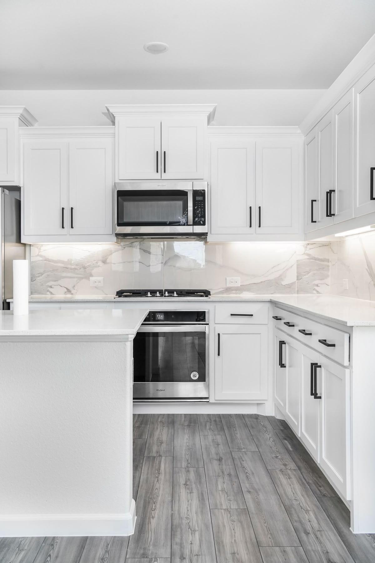 Modern kitchen with light gray cabinetry, white countertops, black fixtures, three wooden stools at island, and a window overlooking greenery.